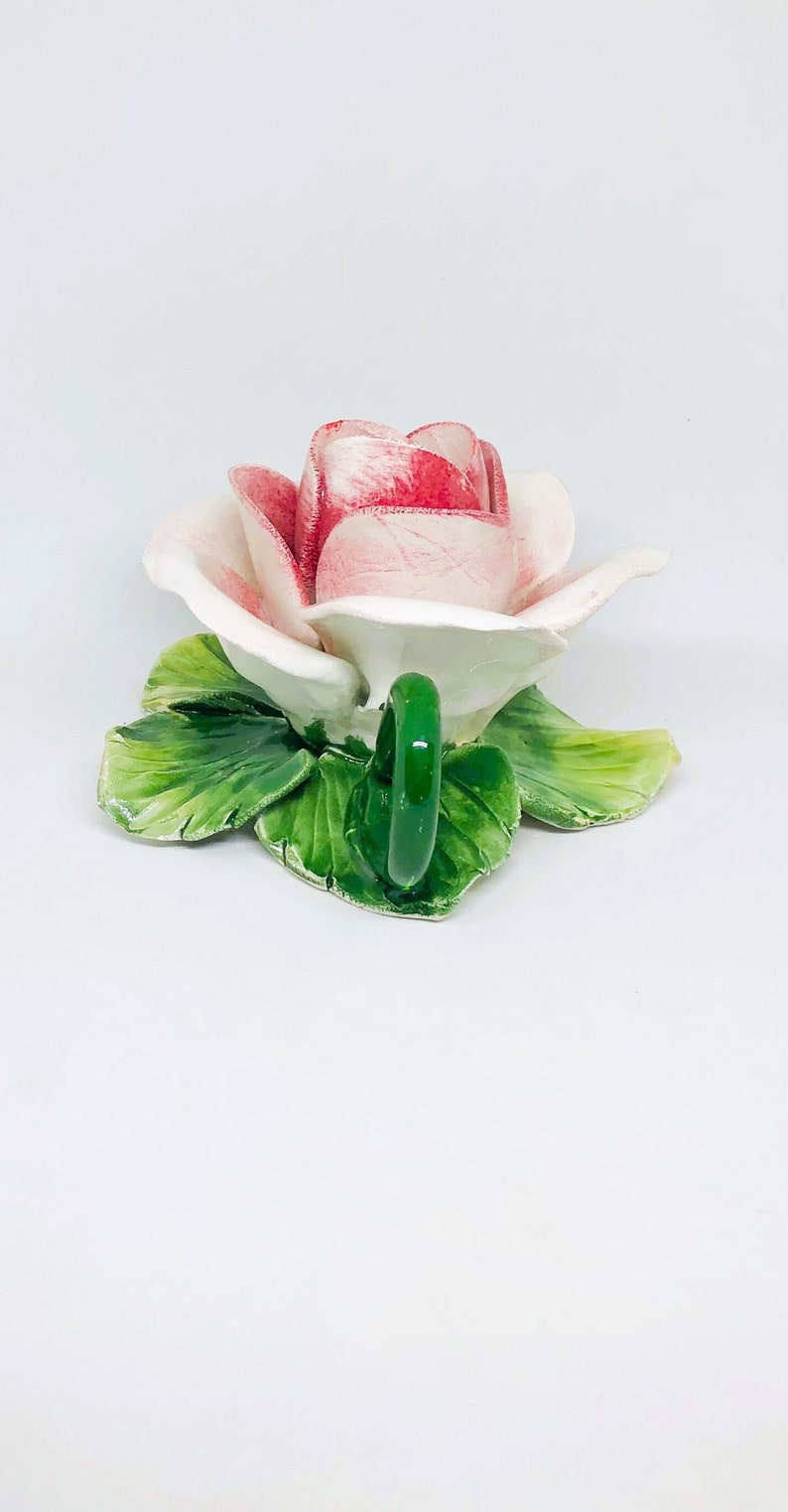 May include: A ceramic rose-shaped teacup with a green handle and green leaves. The rose petals are white with red edges. The teacup is on a white background.