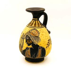 Puede incluir: Un jarrón de cerámica amarillo y negro con asa. El jarrón presenta una silueta negra de una persona con una lira y un borde decorativo. El jarrón mide aproximadamente 18 cm de altura.