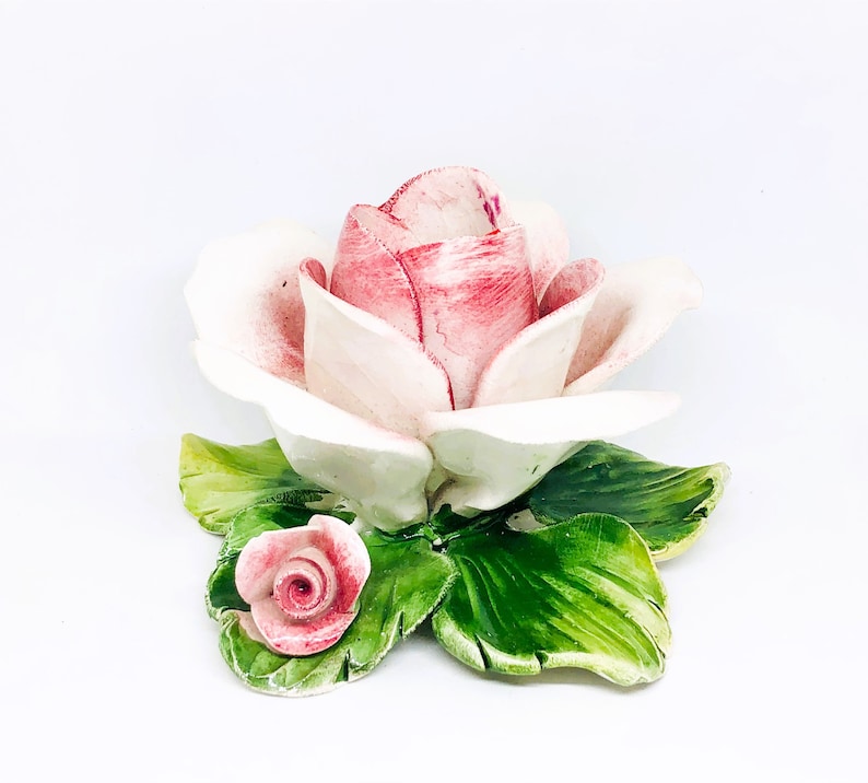 May include: A ceramic rose sculpture with a large, open bloom in shades of pink and white. The petals have delicate veining. The sculpture includes green leaves and a smaller rosebud. The piece is set against a white background.