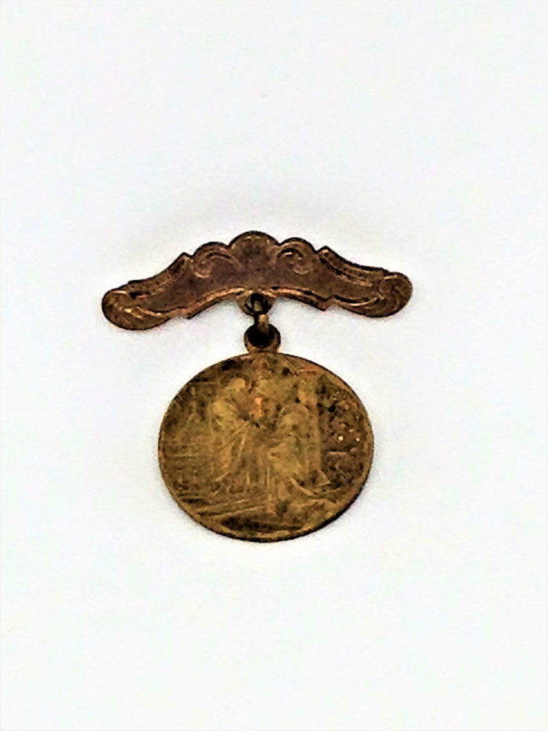 May include: A gold-colored metal pin with a round, detailed design and a curved top. The pin has a decorative, ornate design.