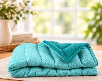 May include: A folded quilted blanket featuring a teal and white geometric diamond pattern on one side and a solid teal plush texture on the reverse. The blanket is rectangular with stitched horizontal channels throughout the fabric.