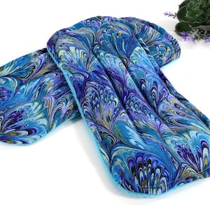 May include: A rectangular fabric heating pad featuring a vibrant marbled pattern in shades of deep blue, turquoise, purple, and black. The pad has a soft, quilted texture with rounded corners and a solid light blue fabric backing on the underside.