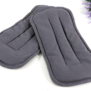 May include: Two gray, rectangular cloth inserts with rounded edges, likely for a baby product. The inserts have a quilted design with vertical stitching. A sprig of purple flowers is in the background.