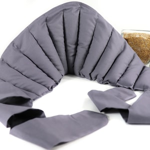 May include: A gray, contoured, weighted neck wrap. The wrap is made of a soft fabric and is designed to drape around the neck and shoulders. The wrap is filled with a weighted material. A glass jar with seeds and a small plant are in the background.