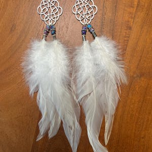 May include: A pair of long dangle earrings featuring silver-toned metal Celtic knot charms at the top. Below the charms are small, round, multi-colored purple and blue beads with silver accents, finished with long, white, soft natural feathers.