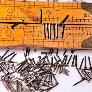 May include: A wooden ruler with a brass end cap and a metal measuring tape. The ruler is labeled "Rabone 9" and "Made in England" and has a number "2550" on the brass end cap. The ruler is surrounded by a pile of small black tacks.