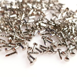 May include: A large pile of silver-coloured tacks or upholstery nails. The tacks are scattered on a white surface.