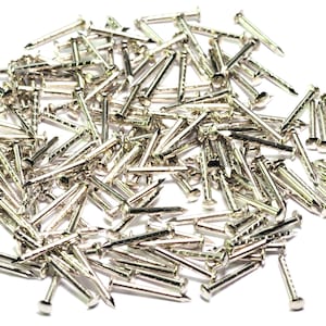 May include: A pile of silver-coloured tacks or brads, a type of nail with a small head, scattered on a white surface.