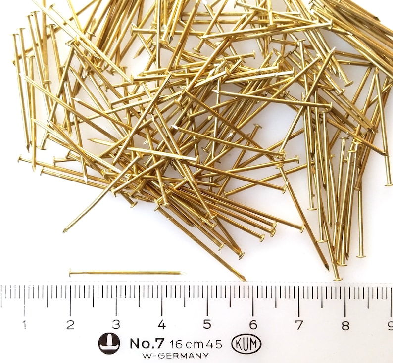 Puede incluir: Un mont&oacute;n de tachuelas o chinchetas doradas con una regla para la escala. La regla muestra medidas en cent&iacute;metros y pulgadas. El texto en la regla dice "No. 7 16 cm 45 KUM W-GERMANY".