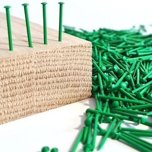 May include: A close-up of a wooden block with four green tacks hammered into it. A large pile of green tacks spills out from the block onto a white surface.