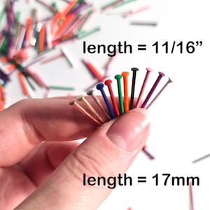 Puede incluir: Primer plano de una mano sosteniendo alfileres de cabeza de colores. Cada alfiler tiene una cabeza de color diferente. La longitud es de 17 mm. Otros alfileres están esparcidos en el fondo.
