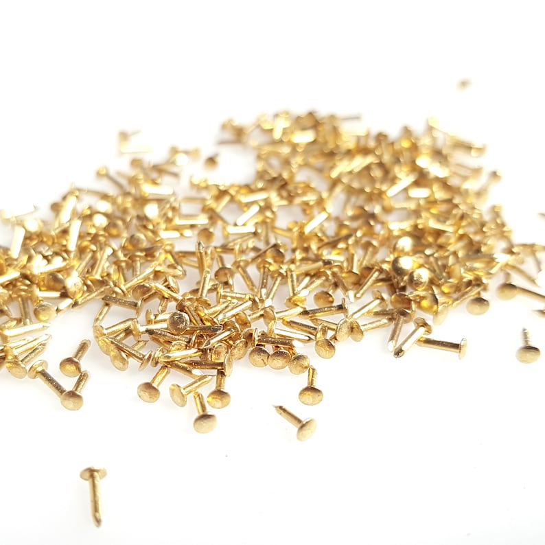 Puede incluir: Un mont&oacute;n grande de tachuelas o clavos de metal de color dorado. Las tachuelas est&aacute;n esparcidas sobre una superficie blanca.