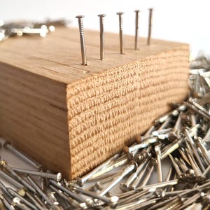 Op de afbeelding: Een houten blok met daarin geslagen verschillende zilveren spijkers. Het blok is omringd door een stapel losse zilveren spijkers.