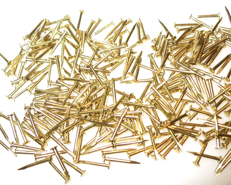 Puede incluir: Un mont&oacute;n grande de tachuelas o clavos de tapicer&iacute;a de color dorado. Las tachuelas est&aacute;n esparcidas sobre una superficie blanca.