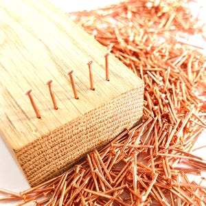 Może przedstawiać: Drewniany blok z rzędami gwoździ miedzianych wbitych w niego. Na pierwszym planie znajduje się duża sterta miedzianych gwoździ.