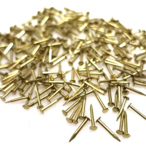 Puede incluir: Un mont&oacute;n grande de tachuelas doradas brillantes o clavos de tapicer&iacute;a. Las tachuelas est&aacute;n esparcidas sobre una superficie blanca.