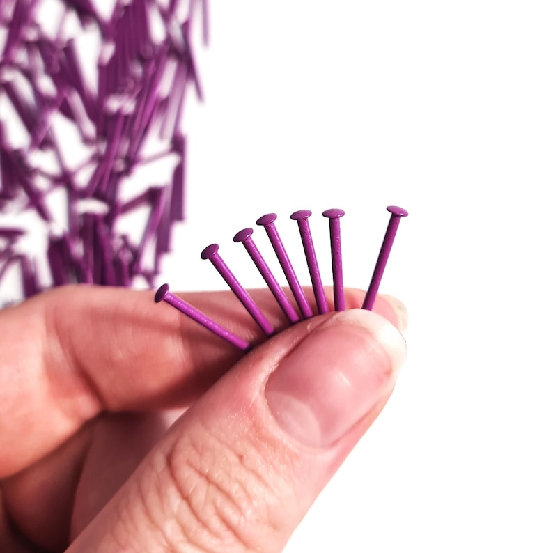 Pode incluir: Close-up de uma m&atilde;o segurando um leque de tachinhas de metal roxas. As tachinhas medem aproximadamente 2,5 cm de comprimento e t&ecirc;m uma cabe&ccedil;a redonda.