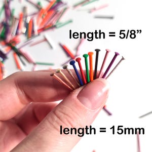 May include: A collection of colorful head pins, each with a different colored head and a silver shaft. The text indicates the length is 5/8 inches or 15mm. The pins are held in a hand against a white background.