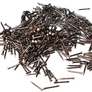 May include: A large pile of antique brass tacks, also known as upholstery tacks, with a shiny finish. The tacks are scattered on a white surface.