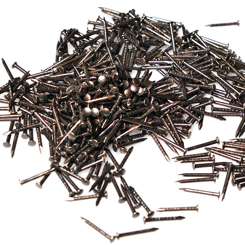 May include: A large pile of antique brass tacks, also known as upholstery tacks, with a shiny finish. The tacks are scattered on a white surface.