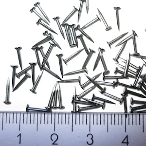 May include: A close-up of a ruler measuring a pile of silver tacks or brads. The ruler measures 10 cm long and the tacks are scattered on a white surface.