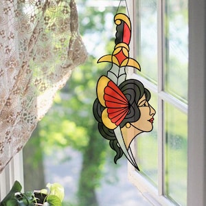 Może przedstawiać: Szklane okno z witrażem wiszące z tradycyjnym wzorem tatuażu przedstawiającym twarz kobiety z sztyletem we włosach. Kobieta ma za uchem czerwony i żółty kwiat i patrzy w prawo. Sztylet jest żółty, czerwony i czarny.