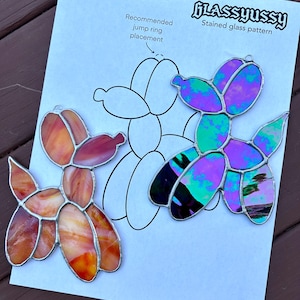 Puede incluir: Dos adornos de perro globo de vidrieras en colores naranja e iridiscente, junto a una hoja de patrones. La hoja tiene un dibujo del perro y el texto "Glassyussy Stained glass pattern" y "Recommended jump ring placement".