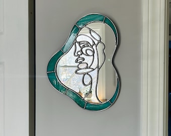 May include: Abstract face-shaped mirror with a teal green border. The mirror features a silver outline of a face. The mirror is mounted on a gray wall. The mirror's shape is irregular, with rounded edges.