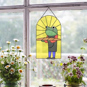 Könnte beinhalten: Ein Buntglasfenster, das in einem Fensterrahmen hängt. Das Buntglas zeigt einen Comic-Frosch, der einen Bäckerhut und eine Schürze trägt und ein Brotlaib hält. Der Hintergrund ist gelb mit Sonnenstrahlen.