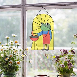 Könnte beinhalten: Ein Buntglasfenster, das in einem Fensterrahmen hängt. Das Fenster zeigt einen Comic-Frosch, der eine rote Schürze trägt und einen Spatel über einem Grill hält. Der Frosch brät eine Wurst. Das Fenster ist gelb mit einem Sonnenstrahlen-Design.