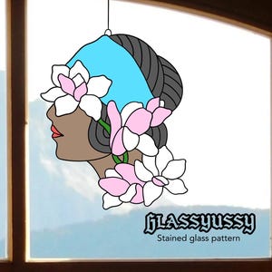 May include: Stained glass pattern featuring a woman's profile with a blue head covering, dark hair, and pink and white flowers. The design includes the text "GLASSYUSSY" and "Stained glass pattern". The artwork is suspended by a thin wire.