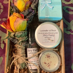 May include: A wooden box filled with a gift set of lavender and rose scented products. The set includes a small blue box with a white ribbon, a small jar of lavender and rose scented lotion, a small bottle of essential oil, and a small jar of rose petals. The box is filled with brown paper shreds.