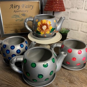 May include: Four silver teapot-shaped planters with different colored polka dots. The planters are on a wooden surface. The planters are decorated with blue, green, pink, and yellow polka dots.