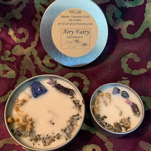 May include: Two silver candle tins with white candles. The larger tin has a blue stone and dried herbs on top of the candle. The smaller tin has dried herbs on top of the candle. The larger tin has a label that reads "Relax Lavender, Chamomile & Basil with Sodalite AiryFairyApothecary.com Airy Fairy Apothecary".