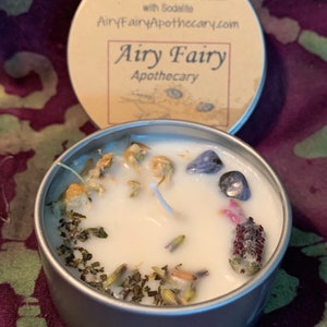 May include: A small, silver tin candle with a white wax base. The candle has a variety of dried herbs and flowers embedded in the wax, including lavender, chamomile, and basil. The tin has a label that reads "Relax Lavender, Chamomile & Basil with Sodalite AiryFairyApothecary.com Airy Fairy Apothecary".