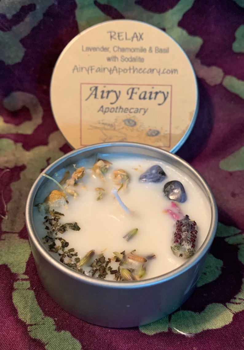 May include: A small, silver tin candle with a white wax base. The candle has a variety of dried herbs and flowers embedded in the wax, including lavender, chamomile, and basil. The tin has a label that reads "Relax Lavender, Chamomile & Basil with Sodalite AiryFairyApothecary.com Airy Fairy Apothecary".
