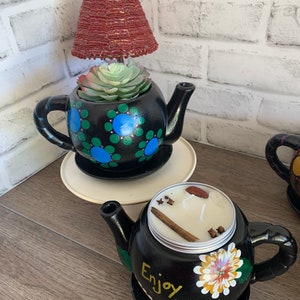 May include: Two black teapots with painted designs. One teapot has a succulent plant inside and a red beaded lampshade on top. The other teapot has a candle inside with cinnamon sticks and cloves. Both teapots have a black and white painted design and the words "Enjoy Life" on the bottom.