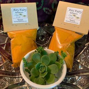 May include: Two brown paper bags filled with yellow turmeric powder. The bags have a label that says "Airy Fairy Apothecary" and "Turmeric". The bags are sitting on a silver tray with a green succulent plant in a white bowl.