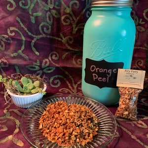 May include: A blue glass jar with a white label that says "Orange Peel" and a small bag of dried orange peel. The jar is sitting on a purple and green patterned fabric.