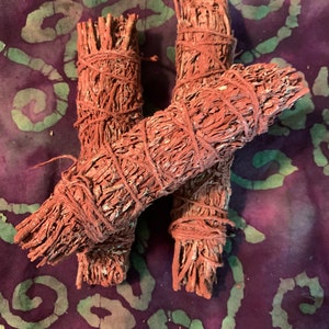 May include: Three bundles of red sage smudge sticks tied together with twine. The sage is a reddish-brown color and is tied together in a bundle.