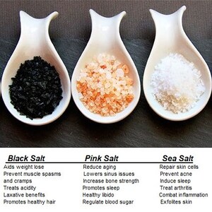 May include: Three white ceramic bowls filled with different types of salt: black salt, pink Himalayan salt, and sea salt. Each bowl has a label with the type of salt and a list of its benefits.