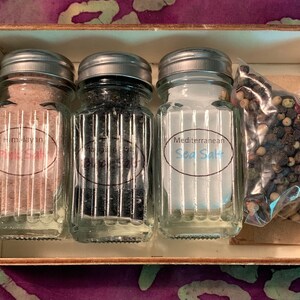 May include: A wooden box with four glass jars containing different types of salt. The jars have silver lids and labels that read "Himalayan Pink Salt", "Black Salt", "Mediterranean Sea Salt", and a bag of peppercorns.