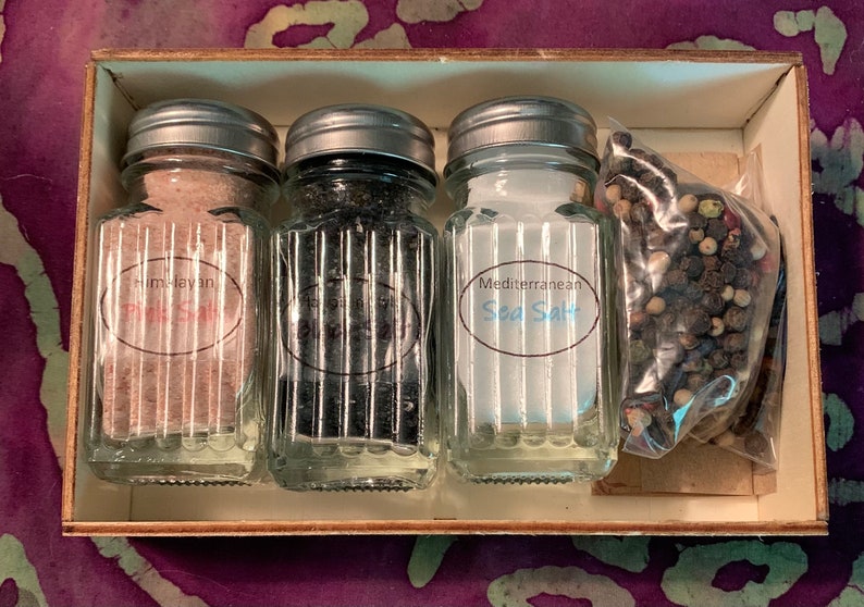 May include: A wooden box with four glass jars containing different types of salt. The jars have silver lids and labels that read "Himalayan Pink Salt", "Black Salt", "Mediterranean Sea Salt", and a bag of peppercorns.