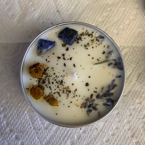 May include: A white candle with dried lavender, chamomile, and green herbs. Blue lapis lazuli stones are embedded in the wax.