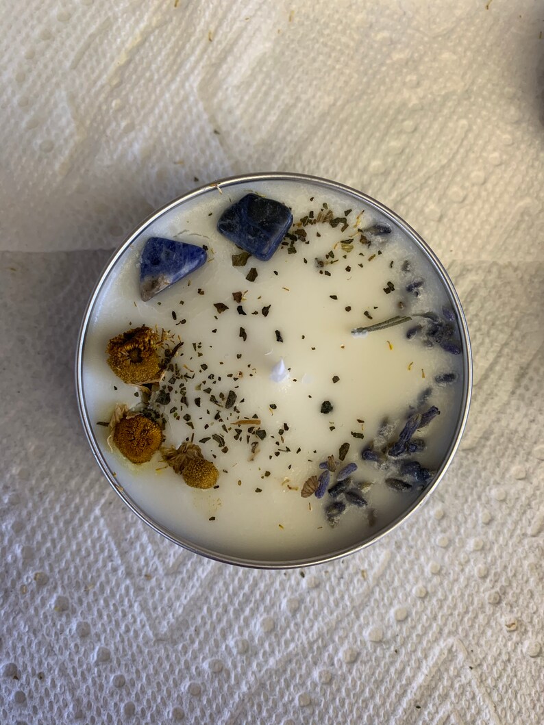 May include: A white candle with dried lavender, chamomile, and green herbs. Blue lapis lazuli stones are embedded in the wax.