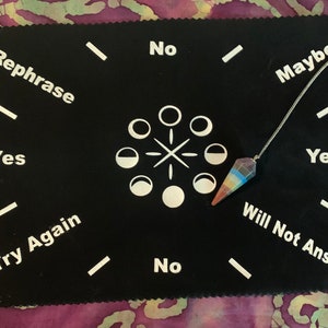 May include: A black felt pendulum board with white text and a circle of moons in the center. The text reads "Rephrase", "Yes", "Try Again", "No", "No", "Maybe", "Yes", and "Will Not Answer". A rainbow crystal pendulum hangs above the board.