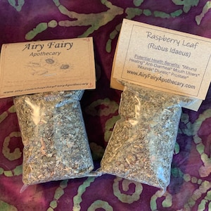 May include: Two small brown paper bags with white text, each containing dried herbs. The bag on the left is labeled "Airy Fairy Apothecary" and the bag on the right is labeled "Raspberry Leaf (Rubus Idaeus)" with a list of potential health benefits.