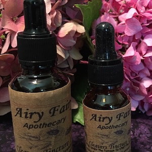 May include: Two brown glass bottles with droppers, labeled "Airy Fairy Apothecary" and "Cleavers Tincture". The bottles are sitting on a purple and white floral fabric.