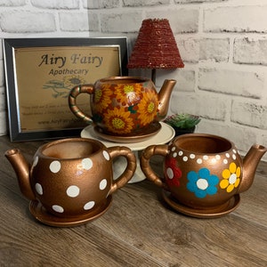 May include: Three decorative teapot-shaped planters with different floral designs. The planters are painted in a copper color with white polka dots, sunflowers, and colorful flowers. The planters are on a wooden surface.