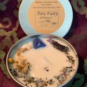 May include: Airy Fairy Apothecary candle in a silver tin with a white wax base. The candle has a blue sodalite crystal, dried lavender, and chamomile flowers embedded in the wax. The label on the tin reads "Relax Lavender, Chamomile & Basil with Sodalite AiryFairyApothecary.com Airy Fairy Apothecary".
